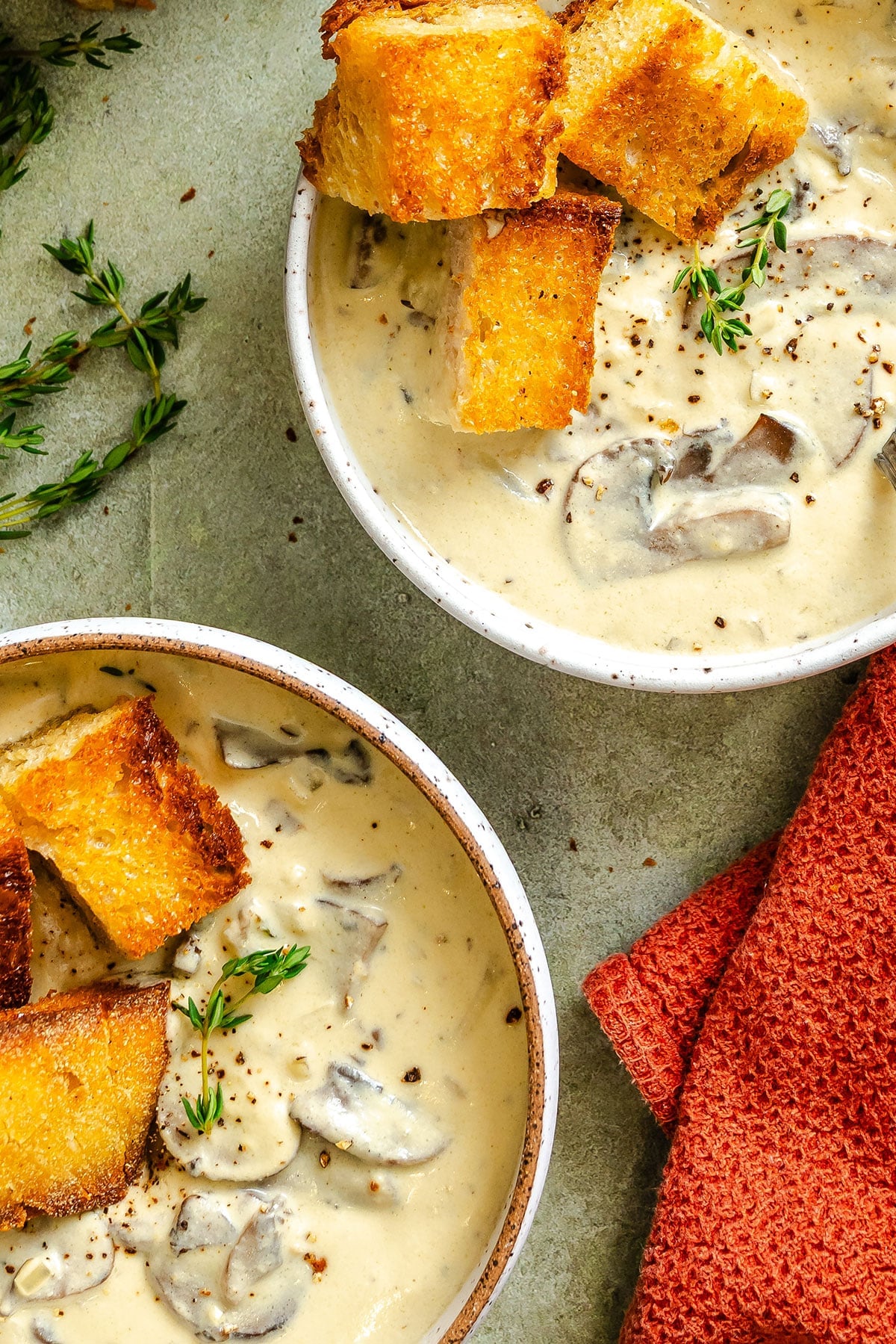 Two bowls of creamy mushroom soup topped with toasted bread cubes and garnished with fresh thyme, placed on a textured surface with a red cloth nearby.