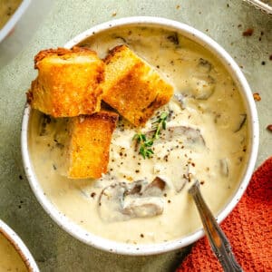 A bowl of creamy mushroom soup garnished with croutons, black pepper, and fresh herbs, with a spoon and an orange cloth napkin beside it.