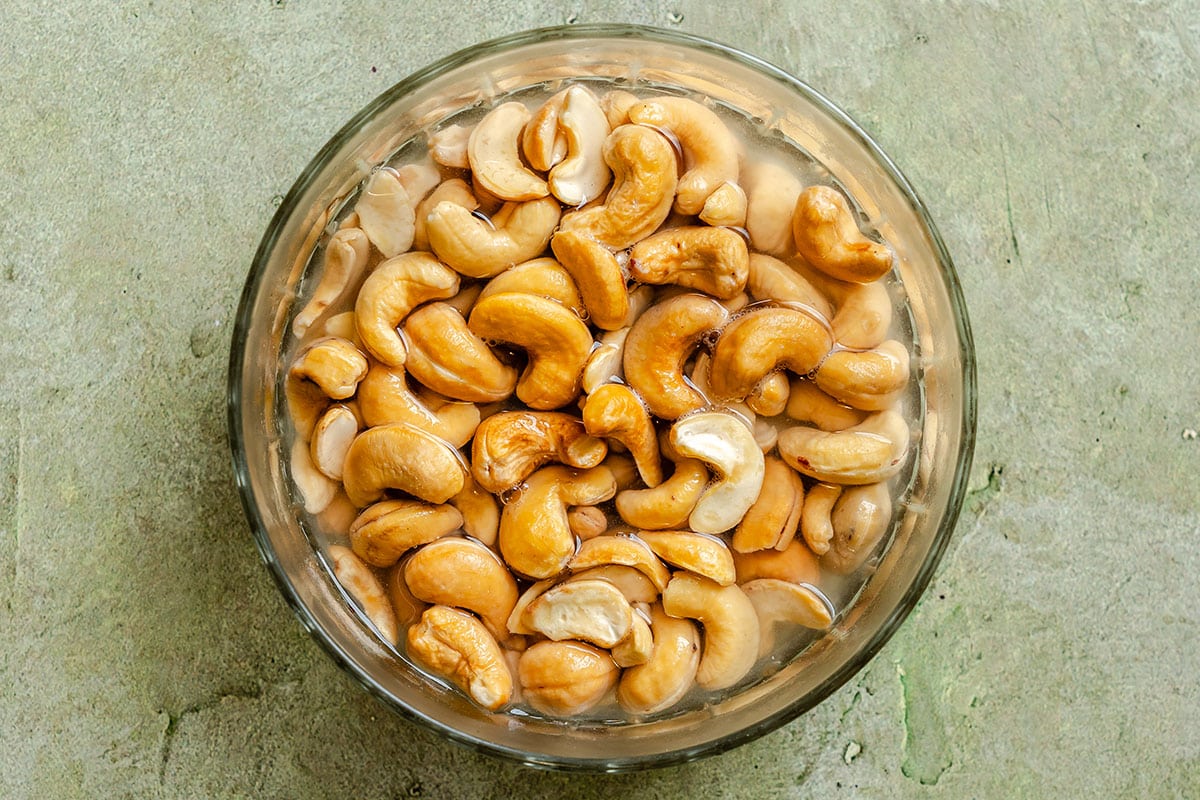 A glass bowl filled with raw cashew nuts soaking in water, placed on a textured light-colored surface.