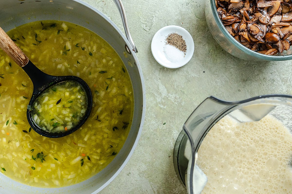 A pot of yellow vegetable soup with a ladle, a small dish of salt and pepper, a bowl of sautéed mushrooms, and a blender with a creamy mixture on a light surface.