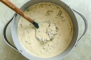 A pot of creamy mushroom soup with visible mushroom slices is being stirred with a wooden spoon on a light-colored surface.