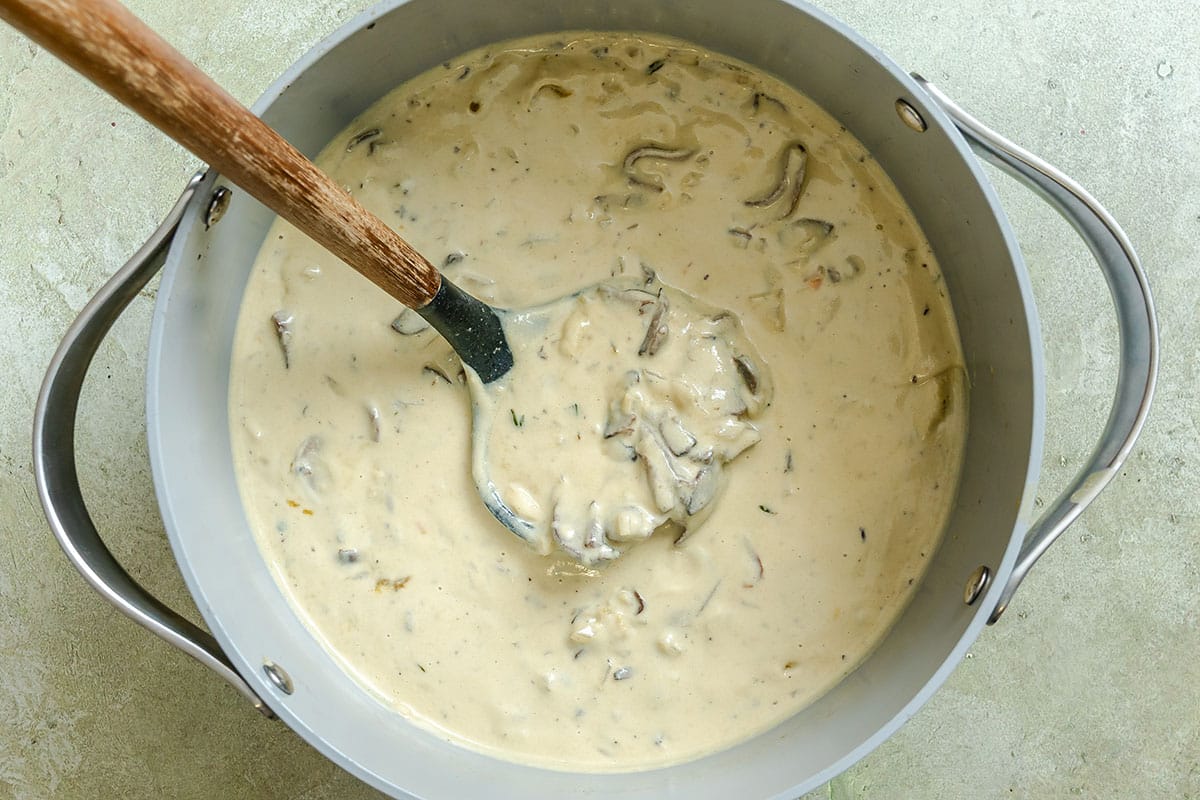 A pot of creamy mushroom soup with visible mushroom slices is being stirred with a wooden spoon on a light-colored surface.