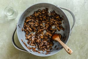 A pan containing sautéed sliced mushrooms with a wooden spoon, placed on a light-colored surface beside an empty glass jar.