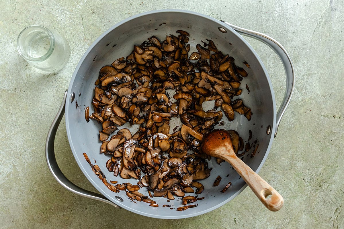 A pan containing sautéed sliced mushrooms with a wooden spoon, placed on a light-colored surface beside an empty glass jar.
