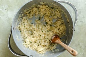 Chopped onions and herbs are sautéing in a large metal pan, with a wooden spoon resting inside.