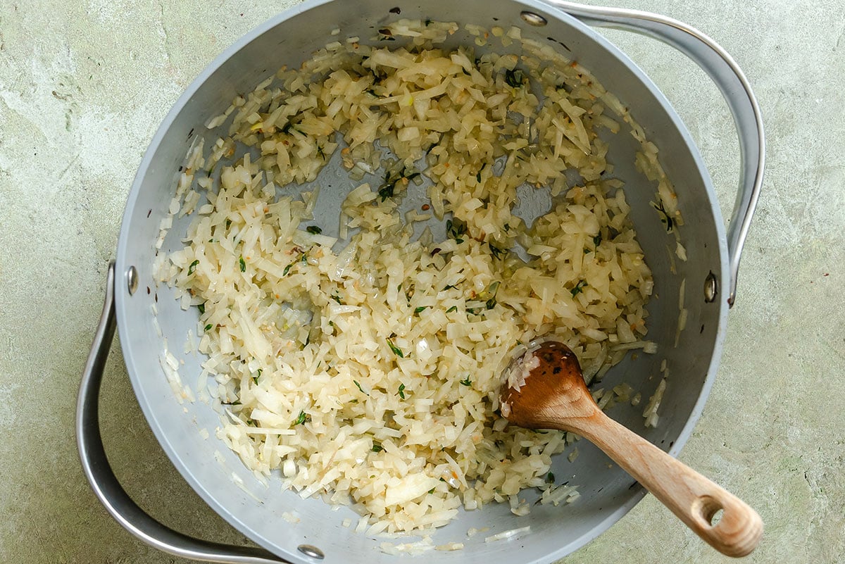 Chopped onions and herbs are sautéing in a large metal pan, with a wooden spoon resting inside.