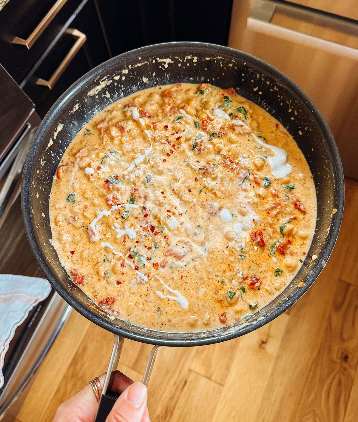 A hand holds a skillet filled with creamy, high-protein chickpea curry—reminiscent of marry me chickpeas—garnished with herbs and coconut milk, set on a wooden kitchen floor.