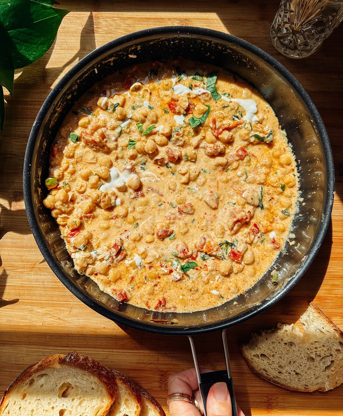 A skillet filled with creamy, high-protein Marry Me Chickpeas and tomato stew, garnished with herbs, sits on a wooden surface next to slices of rustic bread.