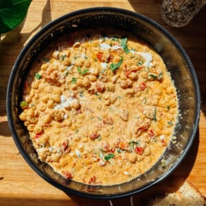 A skillet filled with creamy Marry Me Chickpeas stew, featuring tomatoes, herbs, and a light drizzle of cream, sits on a wooden surface in bright natural light—a high protein delight for any table.