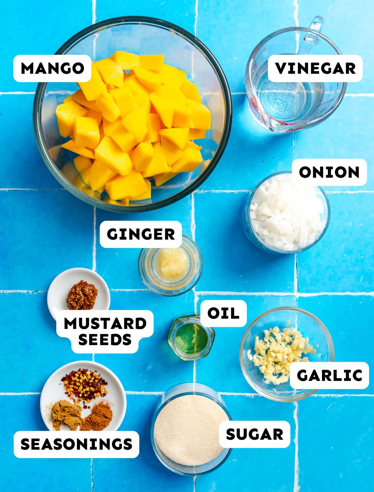 Top-down view of bowls containing chopped mango, vinegar, diced onion, minced ginger, mustard seeds, oil, chopped garlic, sugar, and assorted seasonings, arranged on a blue tiled surface.