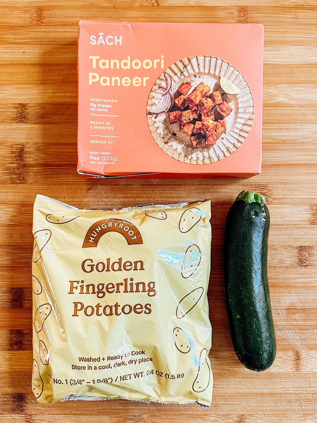 A box of tandoori paneer, a bag of golden fingerling potatoes, and a zucchini are placed on a wooden surface.