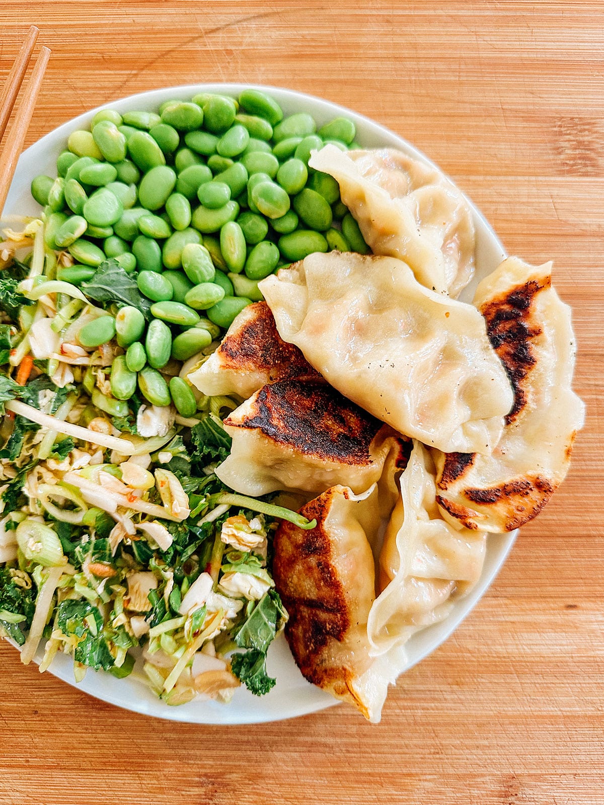 A plate with pan-fried dumplings, a serving of edamame beans, and a mixed green salad on a wooden surface.