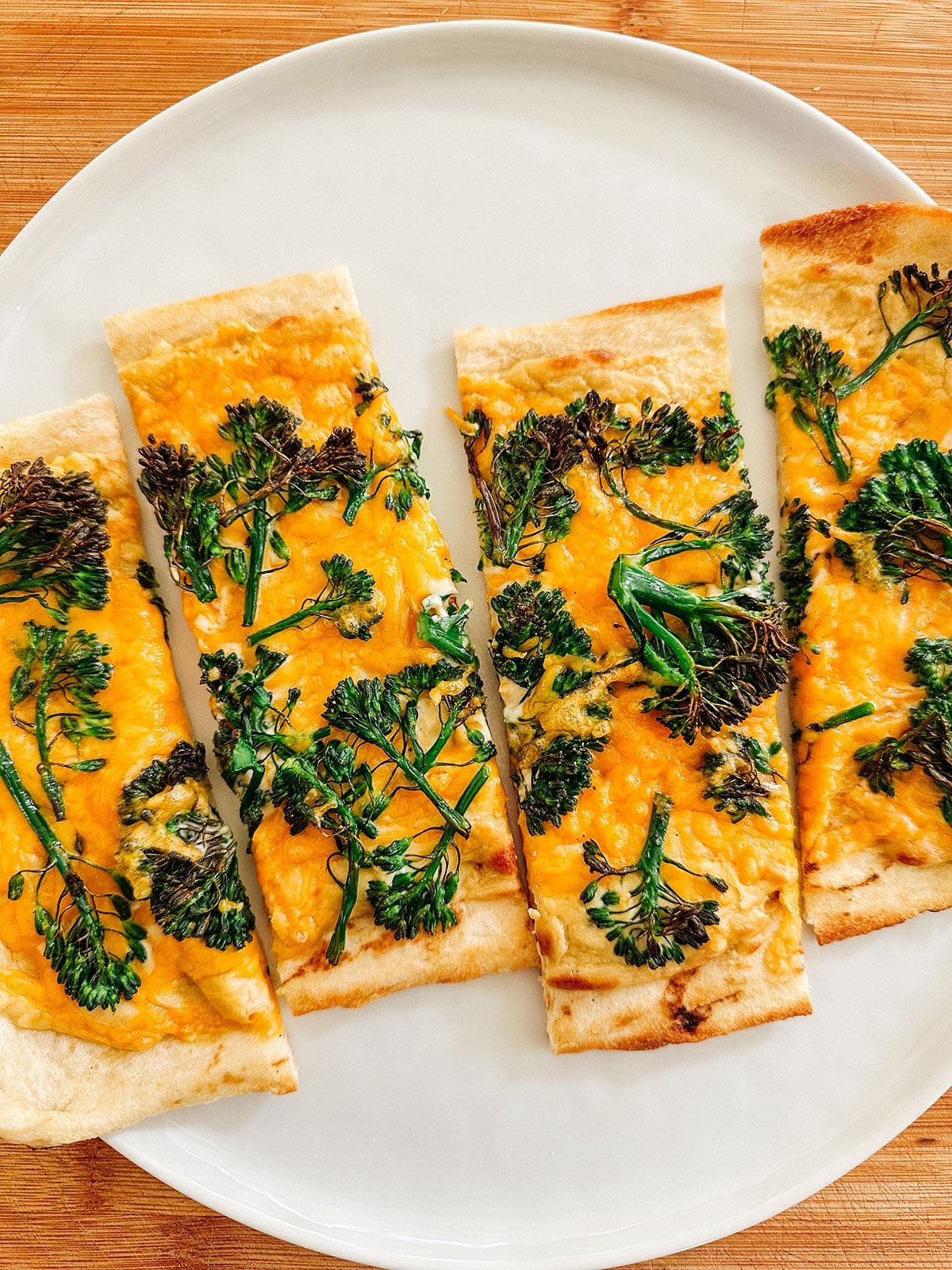 Four rectangular slices of flatbread topped with melted cheese and broccolini are arranged on a white plate.