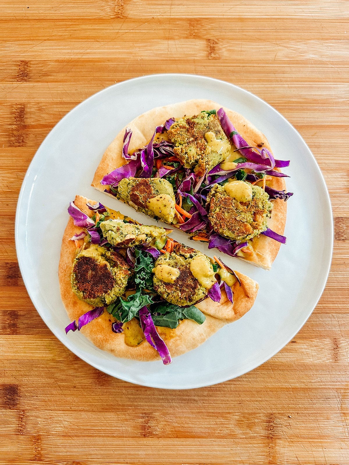 A plate with two pieces of flatbread topped with falafel, sliced red cabbage, greens, and a yellow sauce, on a wooden surface.