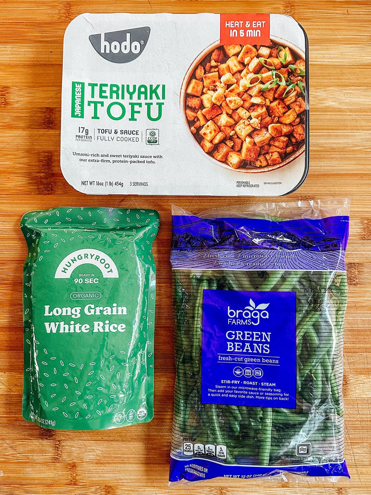 A package of teriyaki tofu, a bag of organic long grain white rice, and a bag of fresh green beans on a wooden surface.