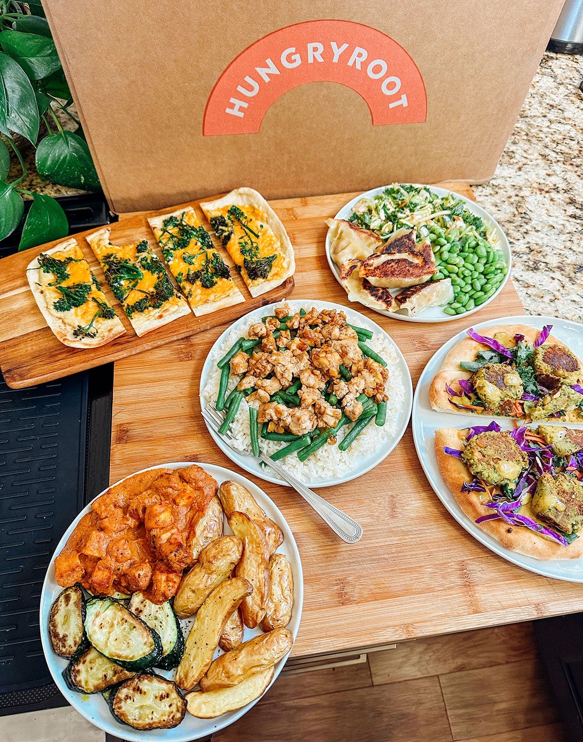 A Hungryroot meal kit box behind a wooden board displaying various prepared dishes, including flatbread, rice bowls, roasted vegetables, and mixed salads.