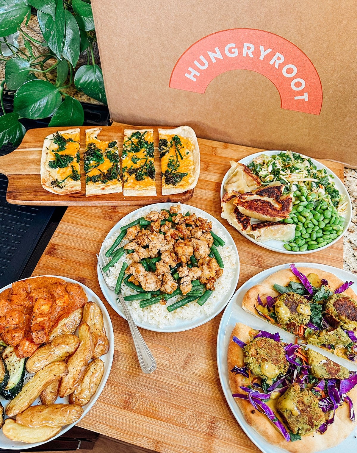 A variety of prepared meals including flatbread, tofu with rice and green beans, salad, potatoes, and a taco plate are arranged on a wooden surface next to a Hungryroot box.