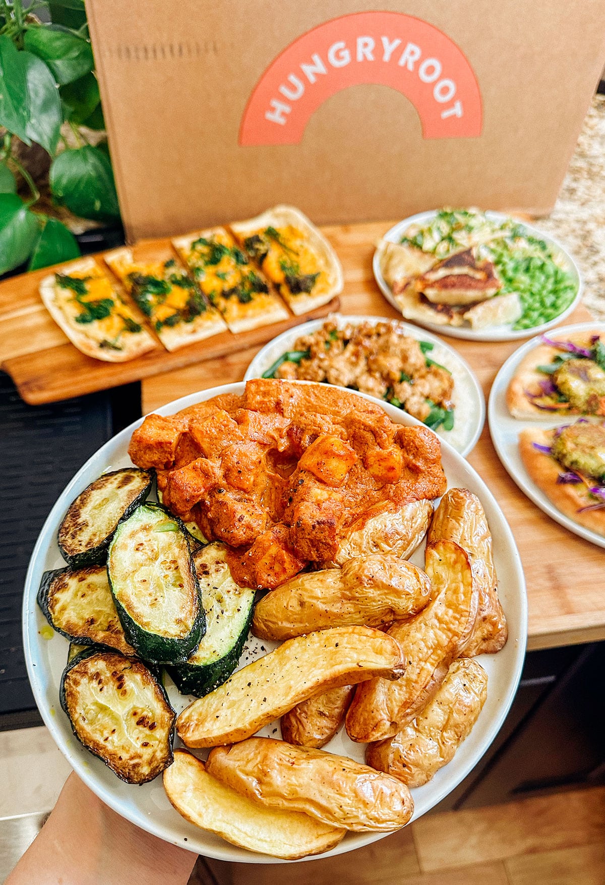 A plate with roasted fingerling potatoes, grilled zucchini, and saucy chicken is held in front of several other vegetable-based dishes and a Hungryroot meal delivery box.