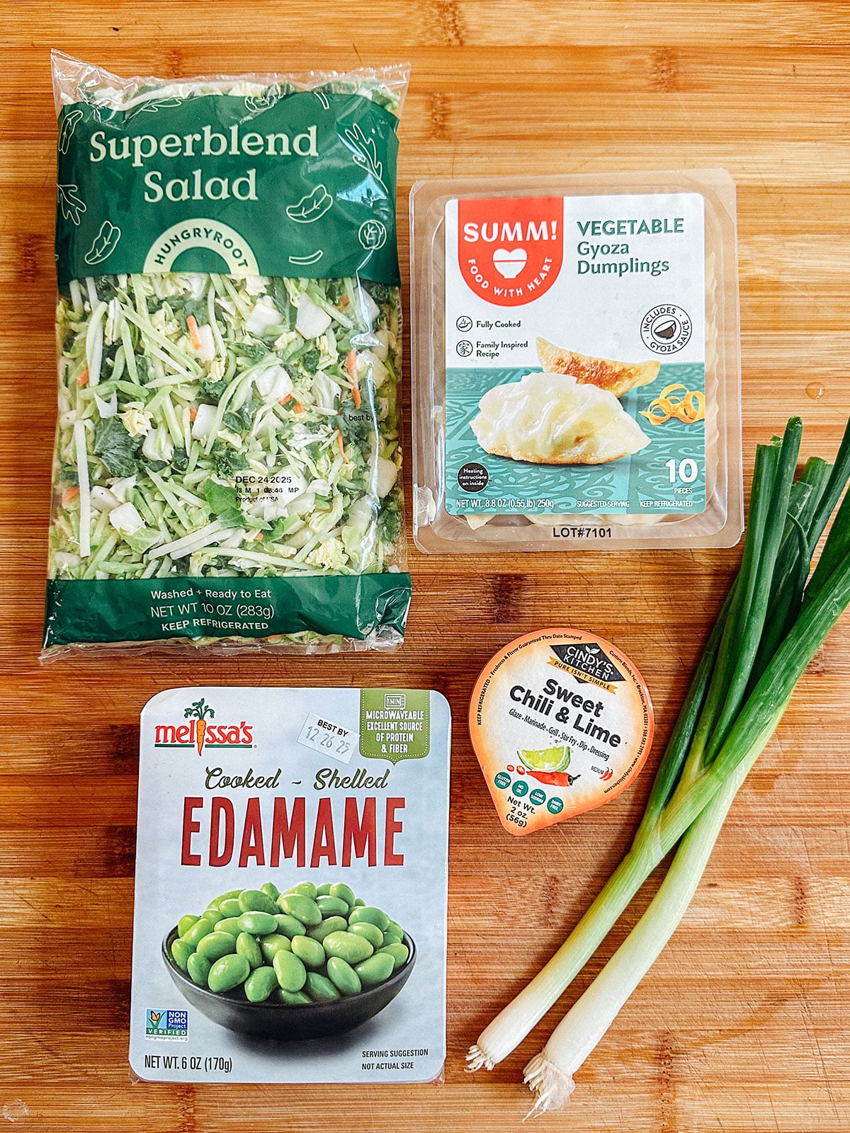A package of superblend salad, vegetable gyoza dumplings, shelled edamame, sweet chili & lime sauce, and two green onions on a wooden surface.