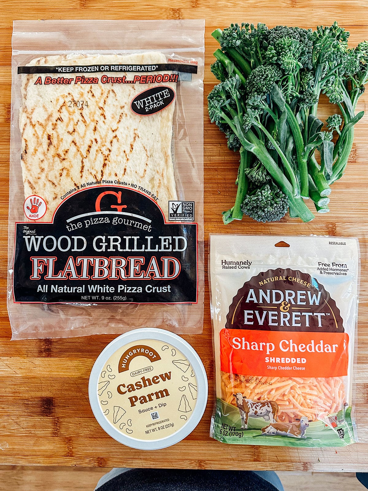 A package of wood grilled flatbread, a bag of shredded sharp cheddar cheese, a container of cashew parmesan, and a bunch of broccolini on a wooden surface.