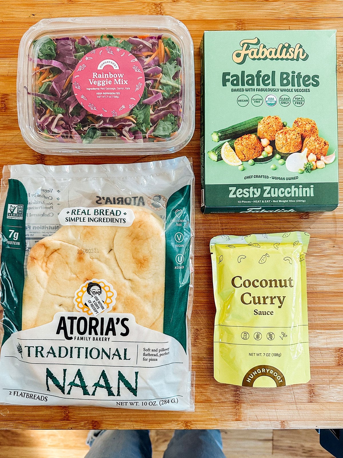 A container of rainbow veggie mix, a box of zucchini falafel bites, a package of traditional naan, and a pouch of coconut curry sauce on a wooden surface.