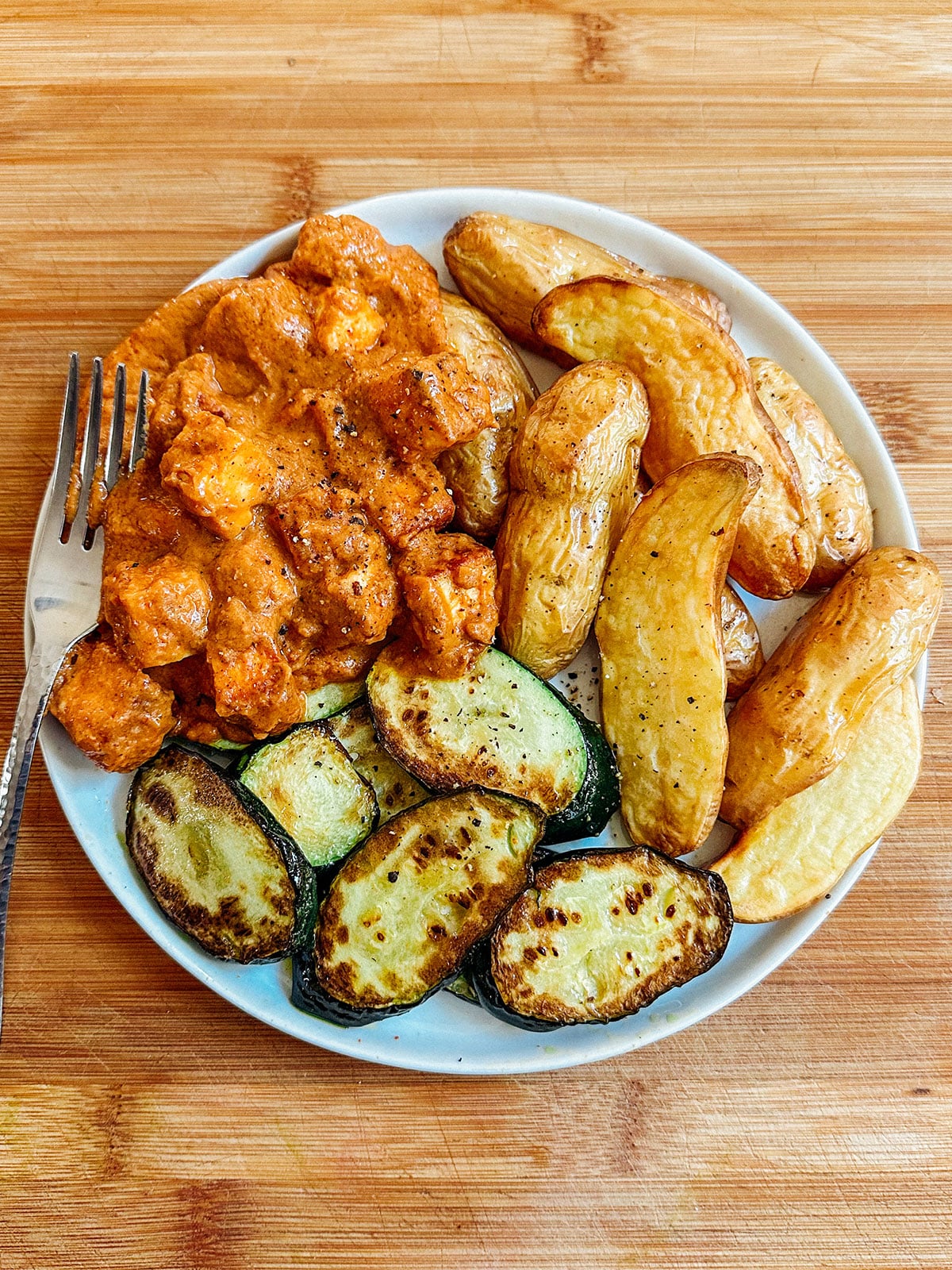A white plate with roasted fingerling potatoes, grilled zucchini slices, and chicken in a creamy orange sauce; a fork rests on the plate.