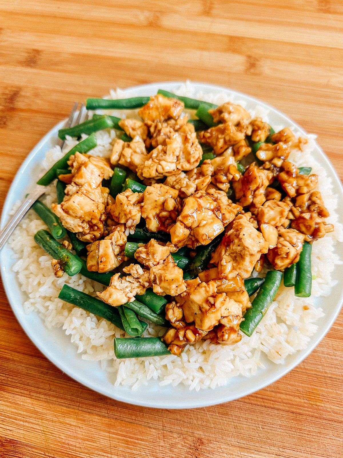 A plate of white rice topped with cooked green beans and diced chicken in a brown sauce, with a fork on the side.