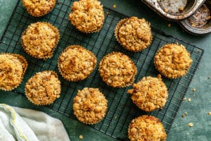 Twelve crumb-topped muffins cool on a black wire rack, with a partially visible muffin tin and striped towel nearby on a green surface.