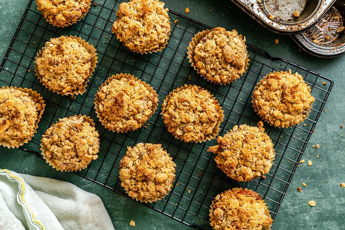 Twelve crumb-topped muffins cool on a black wire rack, with a partially visible muffin tin and striped towel nearby on a green surface.