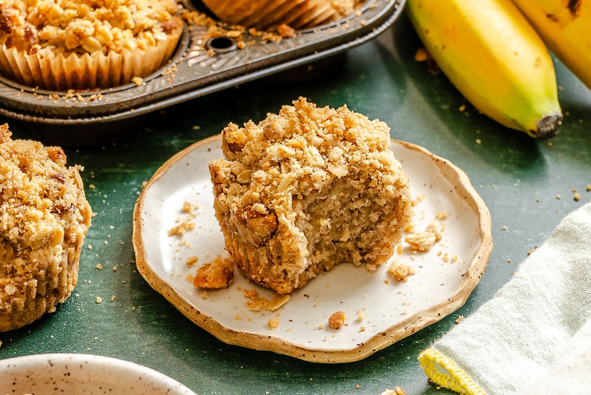 A banana muffin with a crumb topping sits on a plate, partially eaten. More muffins are in a tray nearby, with ripe bananas and a cloth visible in the background.