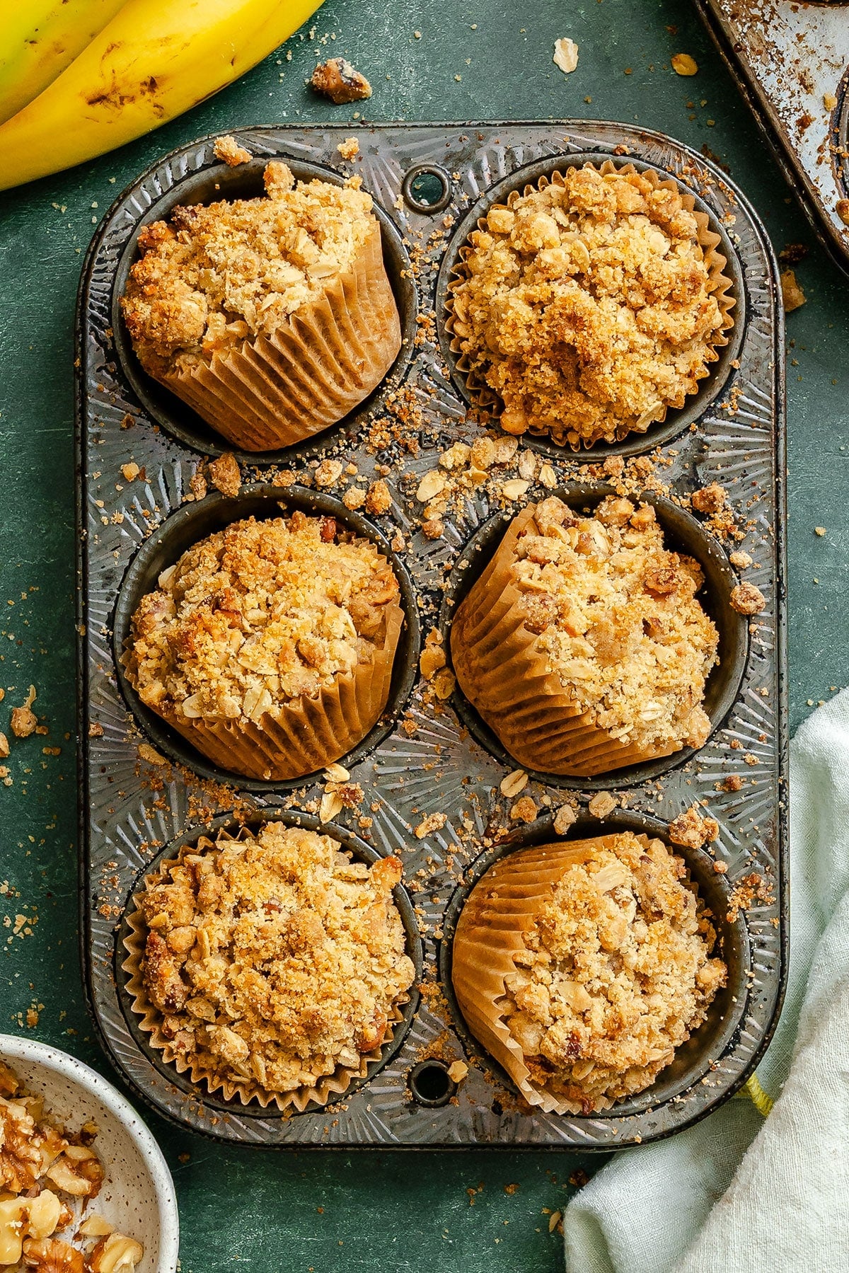 A metal muffin tin holds six banana muffins with crumbly oat streusel topping; some muffins are slightly tilted, and crumbs are scattered around.