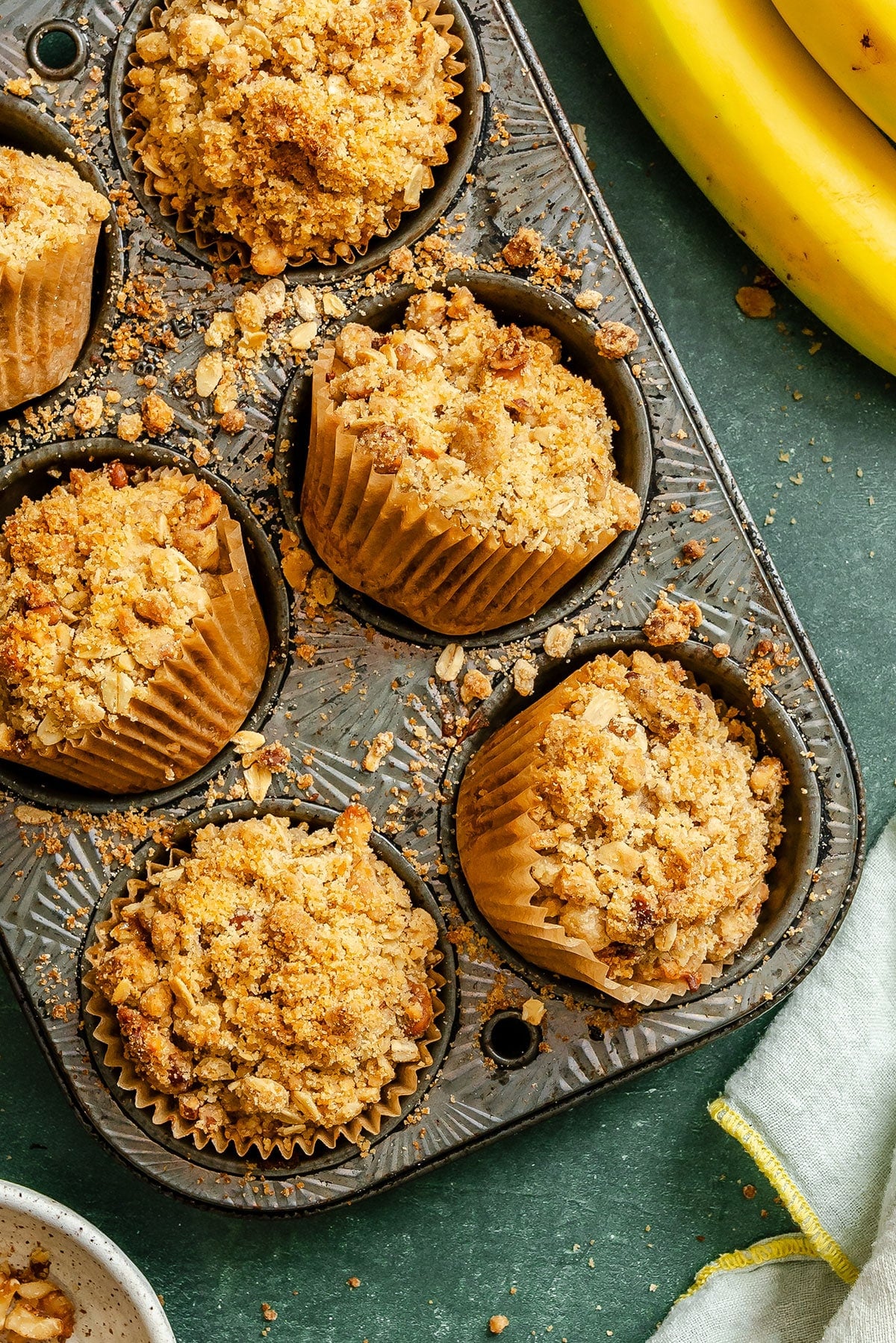 A muffin tin holding six banana muffins with crumb topping, some crumbs scattered around, with bananas and a cloth visible nearby.