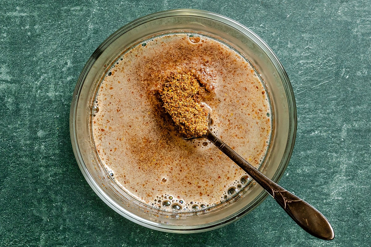 A spoon with ground flaxseed rests over a glass bowl containing a flaxseed and water mixture on a green surface.