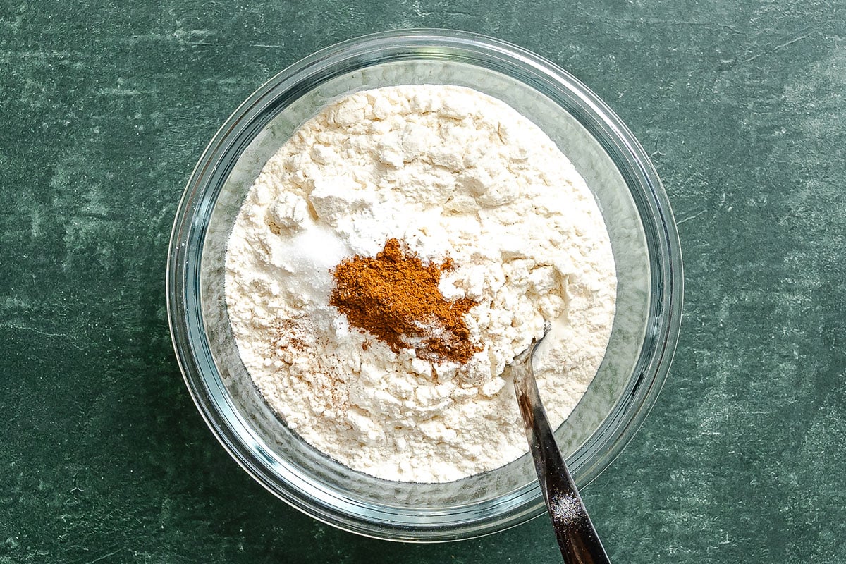 A glass bowl containing flour, cinnamon, and other dry baking ingredients with a metal spoon on a green surface.