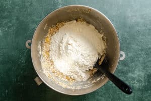 A metal mixing bowl containing flour and other dry ingredients sits on a green surface with a black spatula resting inside.
