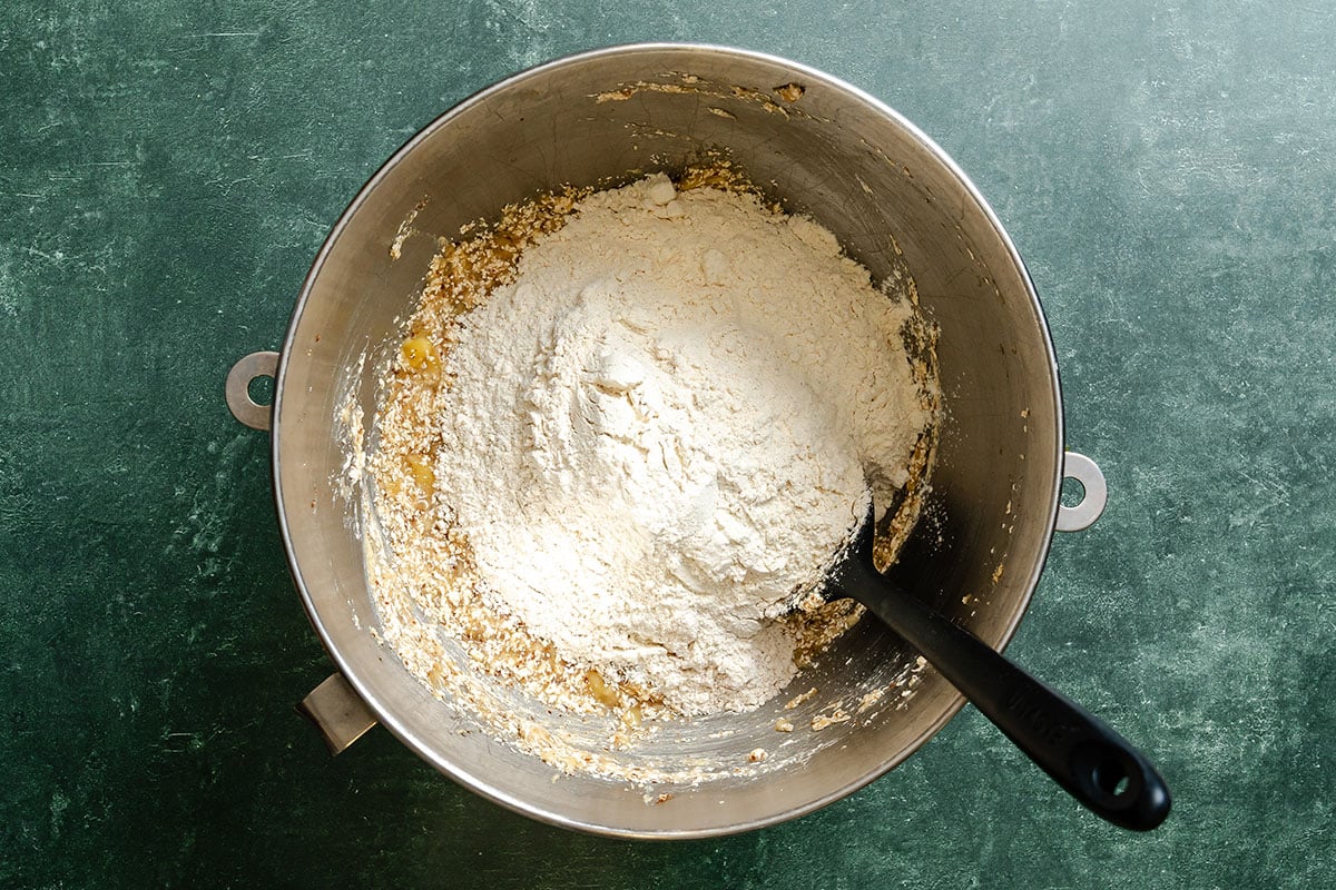 A metal mixing bowl containing flour and other dry ingredients sits on a green surface with a black spatula resting inside.