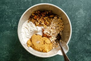 A mixing bowl contains flour, brown sugar, rolled oats, chopped nuts, white sugar, and cinnamon with a fork resting inside.