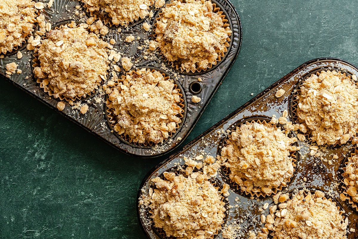 Two muffin pans filled with unbaked muffins topped with oat crumble sit on a dark green surface.