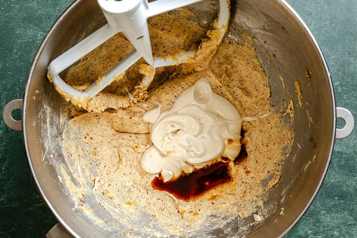 A mixing bowl with a paddle attachment contains partially mixed batter, mayonnaise, and a splash of dark liquid, likely vanilla extract.