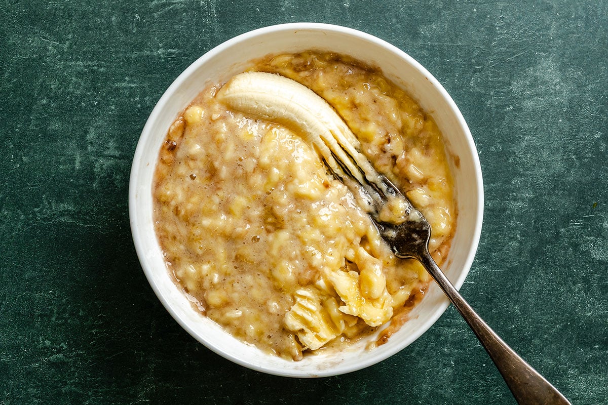 A white bowl with a fork mashing a banana on a green textured surface.