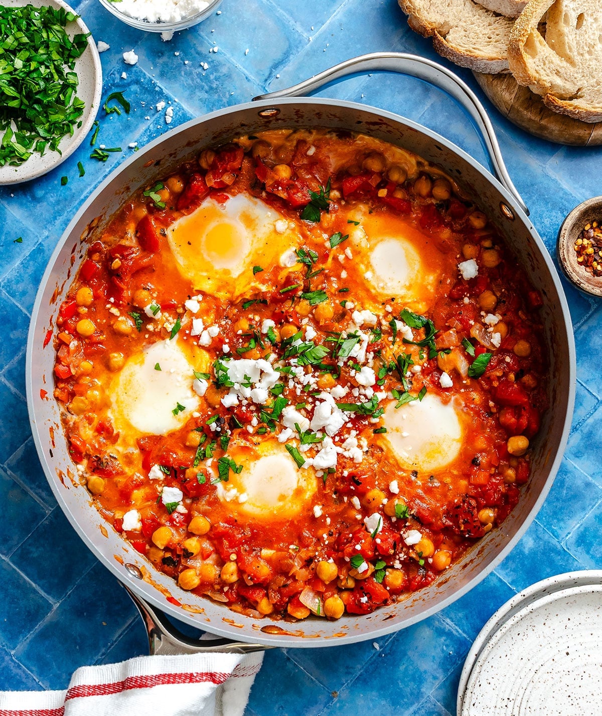 A skillet of shakshuka with poached eggs, tomatoes, chickpeas, herbs, and crumbled cheese, surrounded by bread, parsley, and seasoning bowls on a blue surface.