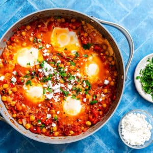 A skillet of shakshuka with poached eggs, chickpeas, tomato sauce, and herbs, topped with crumbled cheese, sits on a blue tile surface next to small bowls of chopped parsley and cheese.
