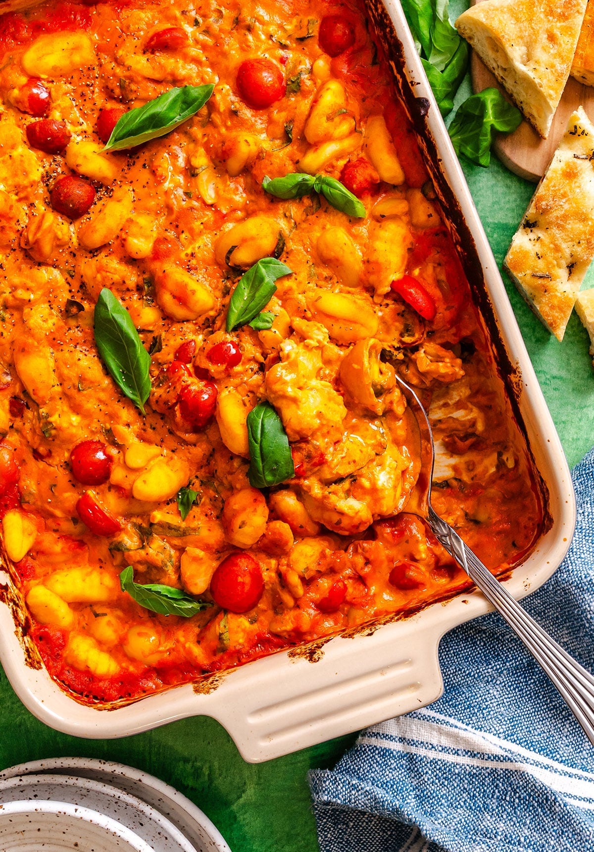 A baked dish of gnocchi in tomato sauce with melted cheese, cherry tomatoes, and fresh basil leaves, served in a rectangular casserole dish with a serving spoon.