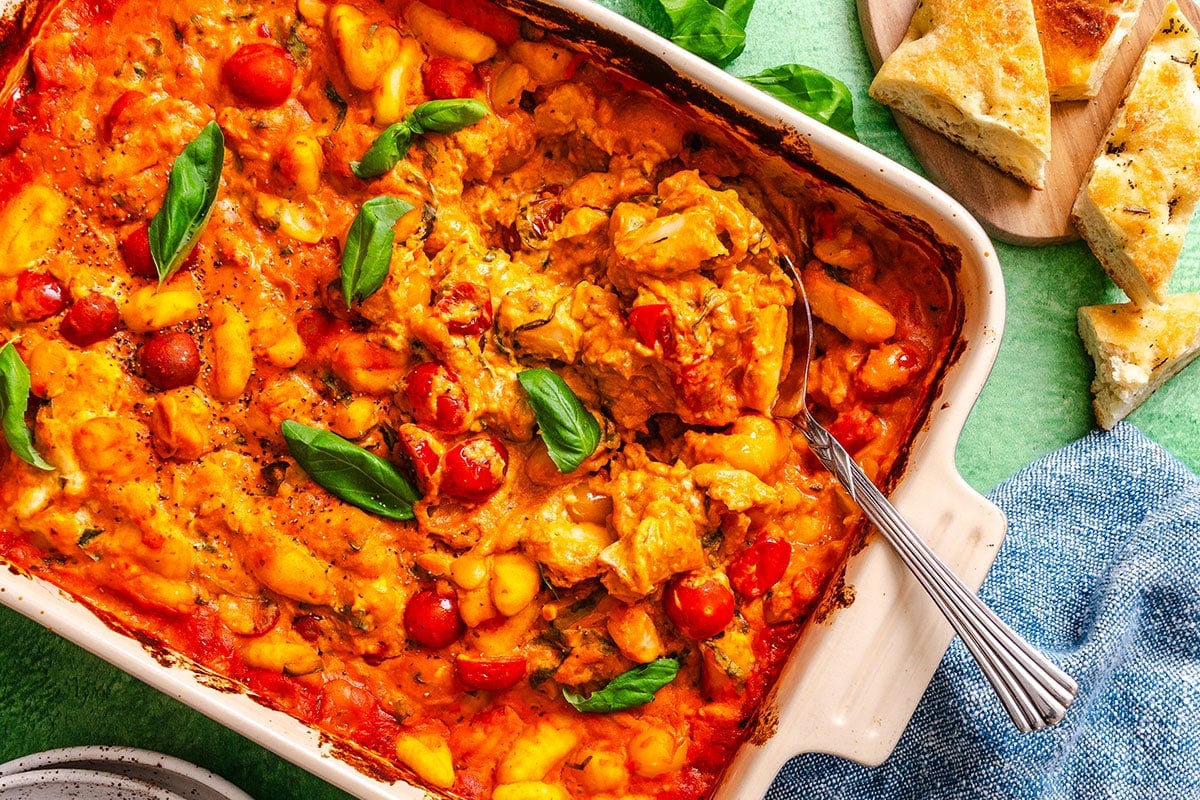 Baked gnocchi with tomato sauce, cheese, chicken pieces, and cherry tomatoes, garnished with fresh basil leaves, served in a white baking dish with bread on the side.