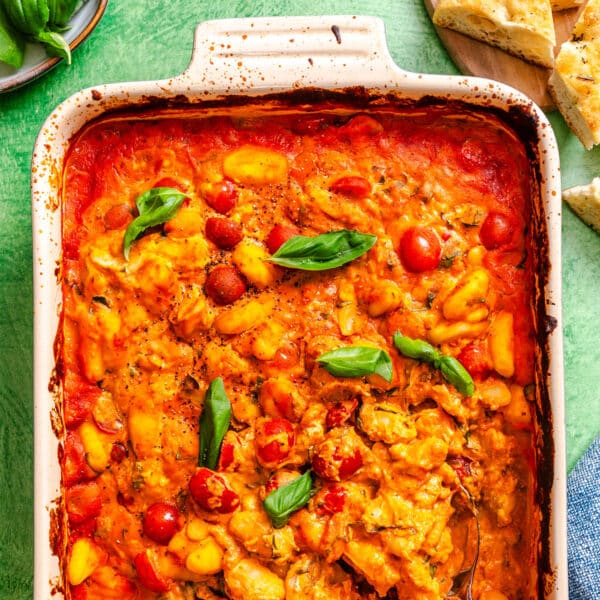 Baked gnocchi in tomato sauce topped with cherry tomatoes and fresh basil leaves in a white casserole dish, with pieces of bread on the side.