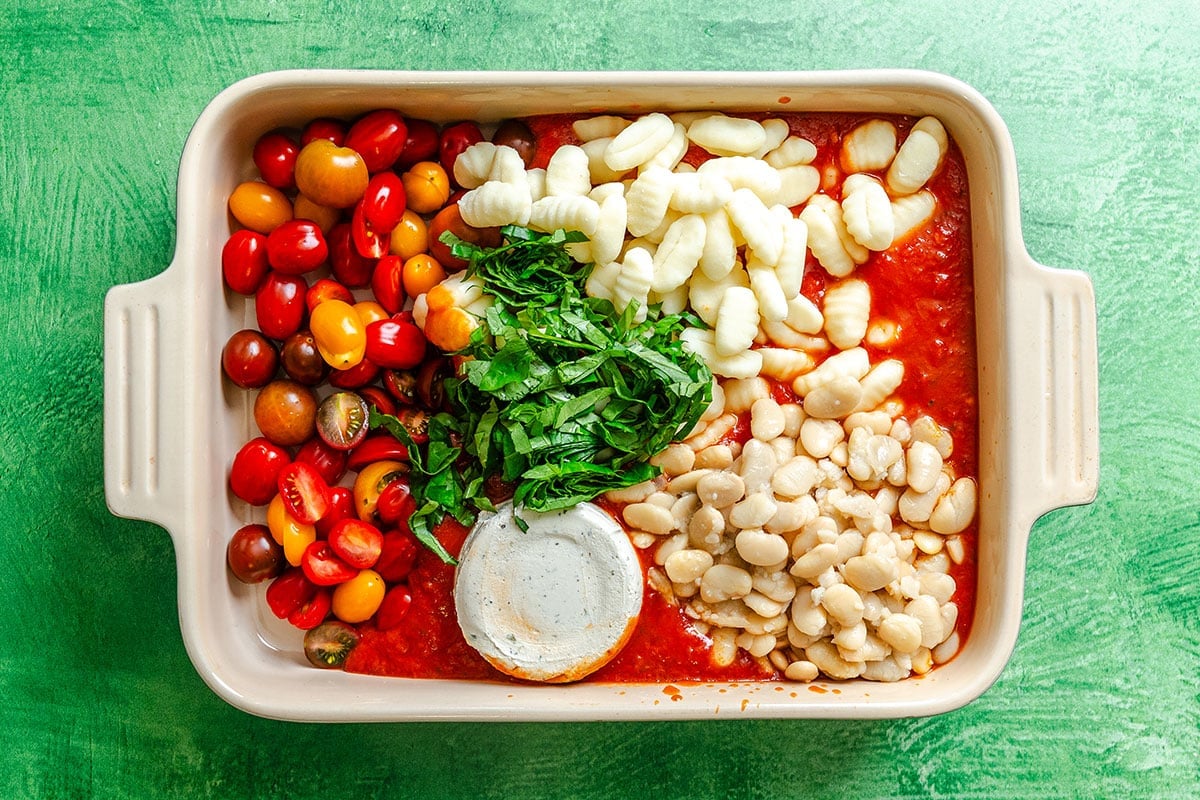 Rectangular baking dish with grape tomatoes, gnocchi, white beans, tomato sauce, spinach, and a round of goat cheese on a green background.