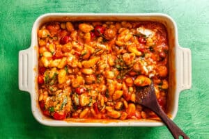 A white baking dish filled with baked gnocchi in tomato sauce with vegetables, topped with herbs, and a wooden spoon resting inside.