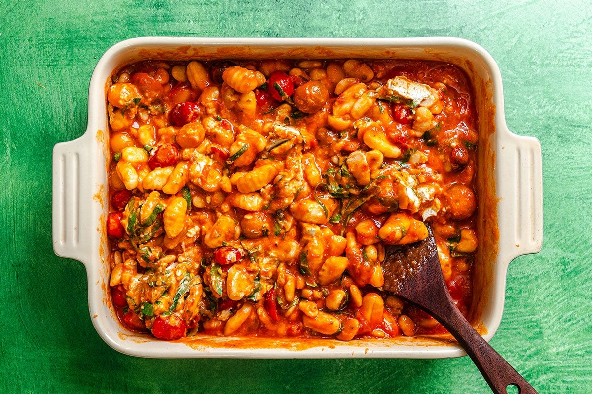 A white baking dish filled with baked gnocchi in tomato sauce with vegetables, topped with herbs, and a wooden spoon resting inside.