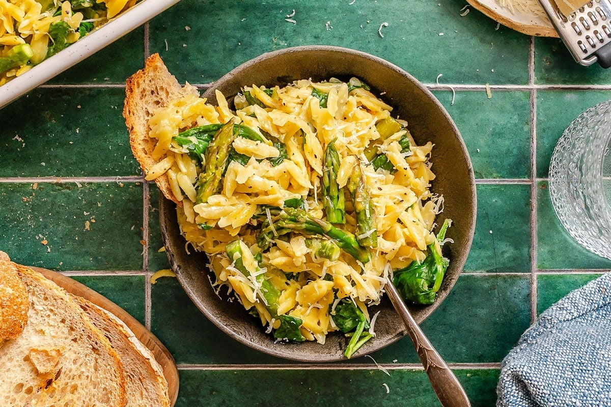 A bowl of orzo pasta with asparagus, spinach, and grated cheese, served with a slice of bread on a green tiled surface.
