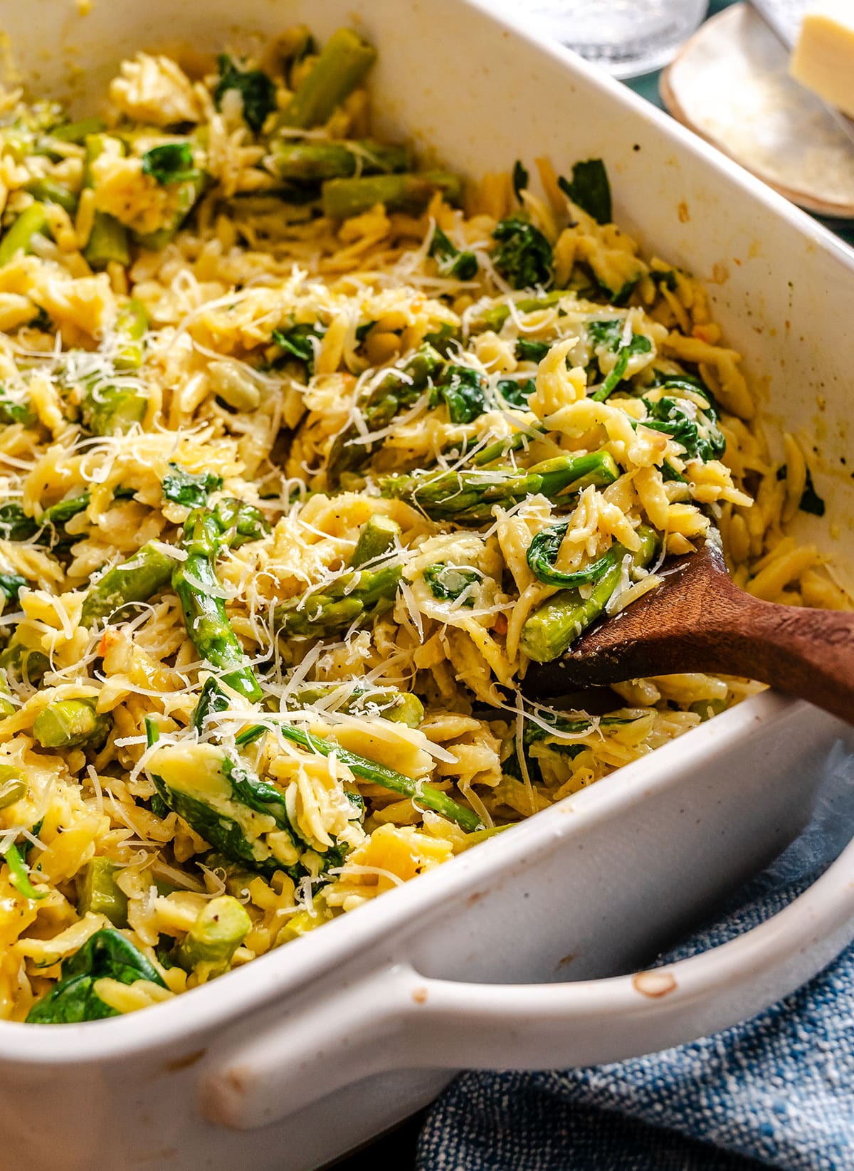 A close-up of baked orzo pasta mixed with asparagus, spinach, and grated cheese in a white ceramic dish, with a wooden spoon on the side.